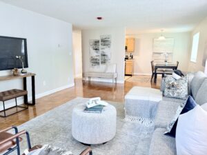 Interior Furnished Unit Living room, Couch and 2 chairs, ottoman in center of room, large area rug, neutral toned walls, tv on far side of wall, abstract art on the walls.