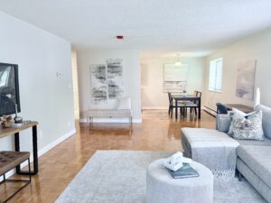 Interior Furnished Unit Living room, Couch and 2 chairs, ottoman in center of room, large area rug, neutral toned walls, tv on far side of wall, abstract art on the walls.