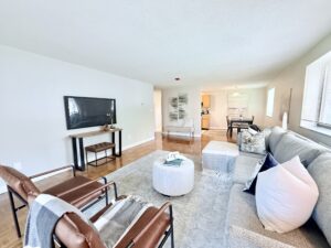Interior Furnished Unit Living room, Couch and 2 chairs, ottoman in center of room, large area rug, neutral toned walls, tv on far side of wall, abstract art on the walls.