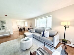 Interior Furnished Unit Living room, Couch and 2 chairs, ottoman in center of room, large area rug, neutral toned walls, tv on far side of wall, abstract art on the walls.