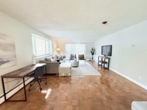 Interior Furnished Unit Living Room, Wood Floors, Workstation on left side of room, couch and 2 chairs in the center of living room, sliding glass doors to balcony.