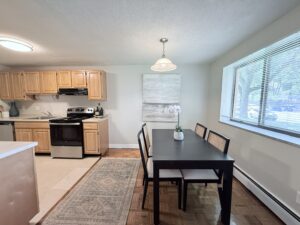 Interior Furnished Kitchen and Dining Area, attached to living room, wood floors, table in dining room, laminate floors in kitchen, light brown wood cabinetry, stainless steel appliances.