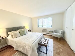 Interior Furnished Unit Bedroom, Wood floors, Neutral toned walls, folding closet doors, window on farside wall.